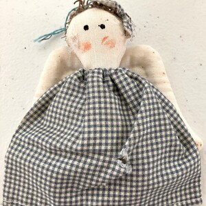 Könnte beinhalten: Eine handgefertigte Stoffpuppe in Engelform mit einem cremefarbenen Körper, aufgenähten Flügeln und einem blau-weiß karierten Kleid. Die Puppe hat ein handgezeichnetes Gesicht mit zwei schwarzen Punktaugen, orangefarbenen Wangen und braunem Garnhaar.