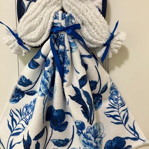 May include: A decorative kitchen towel holder shaped like an angel. The angel's body is a white towel with a blue floral pattern. The angel's wings and hair are made of white terry cloth. Blue satin ribbons are used for accents.