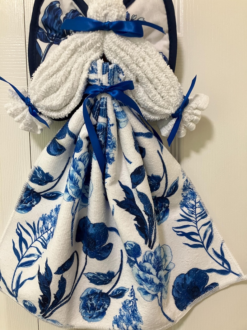 May include: A decorative kitchen towel holder shaped like an angel. The angel's body is a white towel with a blue floral pattern. The angel's wings and hair are made of white terry cloth. Blue satin ribbons are used for accents.