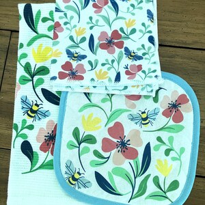 May include: A set of kitchen linens featuring a floral and bee design. The set includes a white textured towel, a square dishcloth, and an oven mitt. The floral pattern includes red, pink, and yellow flowers with green leaves and blue bees.