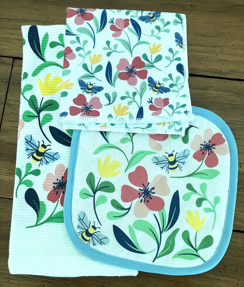 May include: A set of kitchen linens featuring a floral and bee design. The set includes a white textured towel, a square dishcloth, and an oven mitt. The floral pattern includes red, pink, and yellow flowers with green leaves and blue bees.