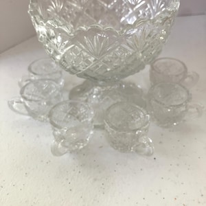 May include: A clear pressed glass punch bowl set featuring a large scalloped-edge bowl with a diamond and fan pattern, accompanied by six matching small glass cups, each displaying the same intricate geometric pressed glass design throughout.