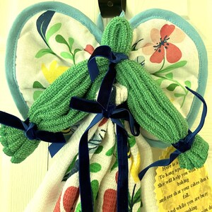 May include: A handmade kitchen angel made from a green washcloth body, floral patterned pot holders for wings, and a patterned kitchen towel. The angel is adorned with navy blue velvet ribbon bows. A yellow tag with text is attached.