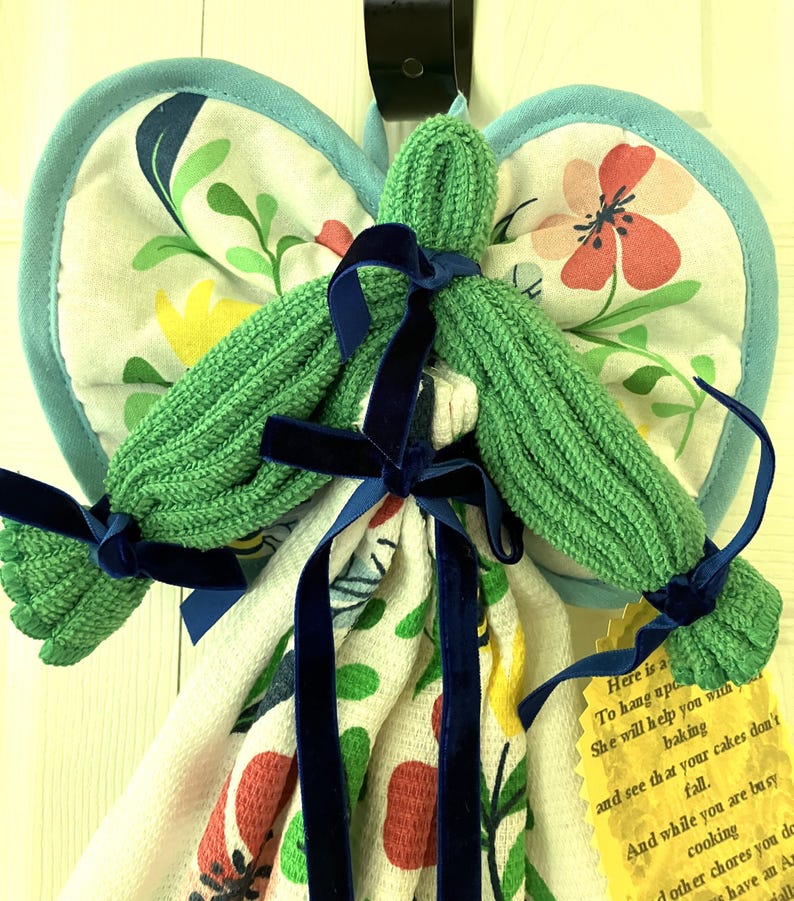 May include: A handmade kitchen angel made from a green washcloth body, floral patterned pot holders for wings, and a patterned kitchen towel. The angel is adorned with navy blue velvet ribbon bows. A yellow tag with text is attached.