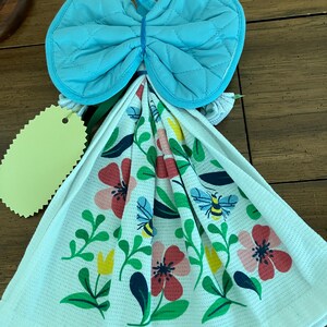 May include: A kitchen towel set with a blue quilted oven mitt tied with a blue rubber band. The white towel features a floral design with red, pink, yellow flowers, green leaves, and bees.