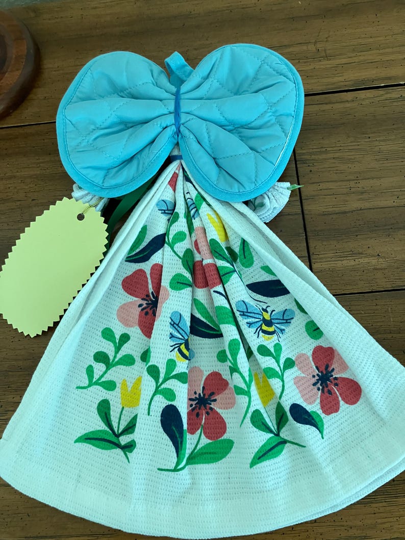 May include: A kitchen towel set with a blue quilted oven mitt tied with a blue rubber band. The white towel features a floral design with red, pink, yellow flowers, green leaves, and bees.