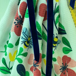 May include: White floral patterned kitchen towel with red, pink, and yellow flowers, green leaves, and blue bees. The towel is accented with a dark blue velvet ribbon.