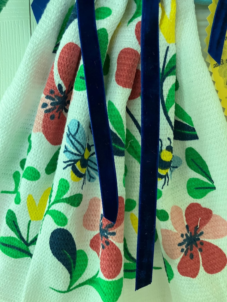 May include: White floral patterned kitchen towel with red, pink, and yellow flowers, green leaves, and blue bees. The towel is accented with a dark blue velvet ribbon.