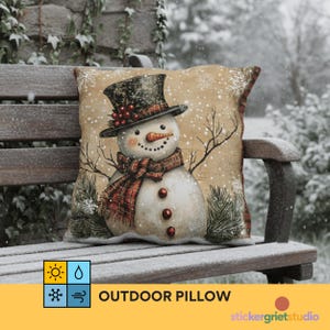 Puede incluir: Un cojín para exteriores con un diseño de muñeco de nieve. El muñeco de nieve lleva un sombrero de copa negro y una bufanda a cuadros rojos y negros. El cojín tiene un fondo beige con nieve y el texto "OUTDOOR PILLOW".