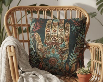 May include: Decorative throw pillow with a detailed hand design in shades of teal, brown, and orange. The pillow is adorned with floral and leaf patterns, resting on a wicker chair with a gray blanket. A small terracotta pot with a succulent is also present.
