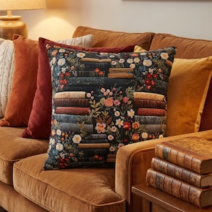 May include: A patterned cushion with a dark backdrop, depicting stacks of books and floral designs in red, white, and yellow. The cushion sits on a brown velvet sofa, alongside other cushions in shades of orange and red.