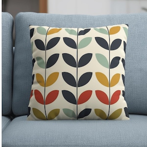 Op de afbeelding: Vierkant sierkussen met een herhalend patroon van gestileerde bladeren in marineblauw, rood, geel en lichtgroen op een crèmekleurige achtergrond. Het kussen ligt op een lichtblauwe bank.