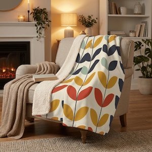 Op de afbeelding: Een crèmekleurige deken met een kleurrijk bladerpatroon in rood, geel, blauw en turquoise. De deken is gedrapeerd over een beige fauteuil met een crèmekleurige sherpa voering. Een tweede gebreide deken is over de arm gedrapeerd.