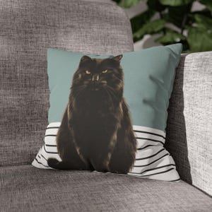 Op de afbeelding: Een vierkant decoratief kussen met een zwarte kat met gele ogen tegen een blauwgroene achtergrond. De onderkant van het kussen heeft een wit en zwart gestreept patroon. Het kussen ligt op een grijze bank.