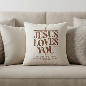 Op de afbeelding: Een crèmekleurig decoratief kussen met de tekst "JESUS LOVES YOU" in bruine letters. Het kussen bevat ook de tekst "WE LOVE EACH OTHER BECAUSE HE LOVED US FIRST 1 JOHN 4:19". Het kussen ligt op een beige bank.