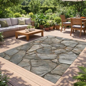 May include: An outdoor rug with a stone pattern in shades of gray and brown. The rug is rectangular and placed on a wooden deck, with outdoor furniture and greenery in the background. The rug adds a natural, textured element to the outdoor space.