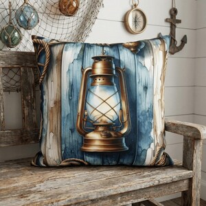 Op de afbeelding: Vierkant decoratief kussen met een vintage messing lantaarn ontwerp tegen een blauw-witte houtnerf achtergrond. Het kussen is afgewerkt met touw en ligt op een houten bank, wat een nautisch thema oproept.