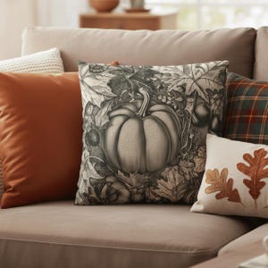 Op de afbeelding: Een verzameling decoratieve sierkussens in herfstkleuren. Het centrale kussen heeft een gedetailleerde grijswaarden illustratie van een pompoen omringd door bladeren. Andere kussens zijn effen oranje, geruit en een crèmekleurig kussen met geborduurde bladeren.