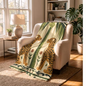 May include: A cream-colored throw blanket featuring a stylized illustration of two cheetahs with brown spots, a green palm tree, and red flowers. The blanket is draped over a cream-colored armchair, creating a cozy and decorative accent.