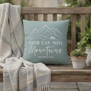Op de afbeelding: Een vierkant, saliegroen kussen met witte tekst en een bergafbeelding. De tekst luidt "FAITH CAN MOVE Mountains" met "MATTHEW 17:20" eronder. Het kussen ligt op een houten bank met een opgevouwen deken.
