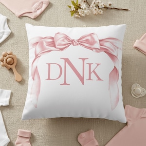 Op de afbeelding: Wit vierkant kussen met een roze strik en de letters "DNK" in roze. Het kussen is omgeven door babyartikelen, waaronder een rammelaar, rompertjes, sokken en een speen. De items zijn in roze en witte tinten.