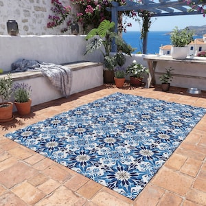 May include: A rectangular outdoor rug with a blue and white floral pattern. The rug is placed on a terracotta tile patio, with a view of the ocean and a white wall with plants and flowers in the background.