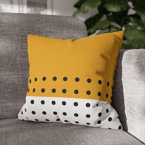 Op de afbeelding: Mosterdgeel en wit decoratief kussen met een stippenpatroon. De bovenste helft van het kussen is effen mosterdgeel, terwijl de onderste helft wit is met gelijkmatig verdeelde zwarte stippen. Het vierkante kussen ligt op een grijze bank.