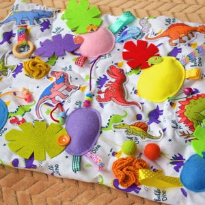 Puede incluir: Una manta de juego blanca para bebés con varias formas de fieltro de colores, incluyendo dinosaurios, hojas y flores. La manta también incluye un anillo de madera, cuentas y cintas.