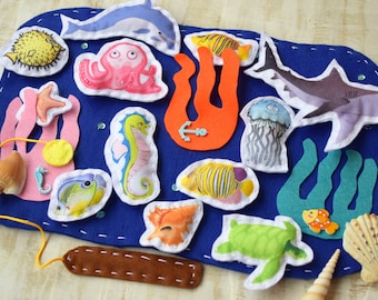 Puede incluir: Un juego de fieltro con temática oceánica que presenta varias criaturas marinas, como un delfín, un pulpo, un tiburón y un caballito de mar, sobre un fondo azul. Incluye una concha y una pieza rectangular marrón.