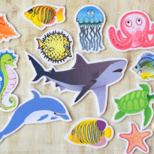 Könnte beinhalten: Ein Set aus bunten Filz-Meerestier-Ausschnitten, darunter ein Seepferdchen, ein Hai, ein Delfin, eine Qualle, ein Tintenfisch, ein Seestern, eine Meeresschildkröte und mehrere Fische. Die Ausschnitte sind perfekt für ein Bastelprojekt für Kinder oder zum Dekorieren eines Raumes.