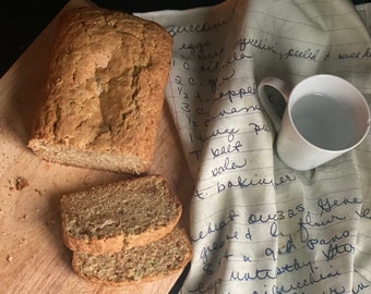 May include: A loaf of sliced courgette bread on a wooden chopping board. A white mug with water sits next to a linen tea towel with handwritten recipe notes. The bread is light brown with a textured crust.