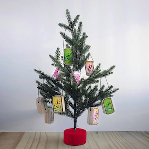 May include: A small artificial Christmas tree decorated with La Croix can ornaments. The ornaments are rectangular with colorful designs and the La Croix logo. The tree has a red base and is set against a white background.