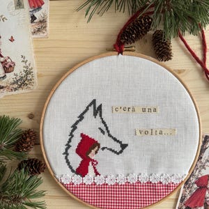 Può includere: Un telaio da ricamo circolare in legno contenente tessuto bianco con un disegno a punto croce di un lupo e una figura con cappuccio rosso. Il testo c'era una volta è ricamato su strisce di tessuto. Un bordo in tessuto a quadretti rossi e bianchi.