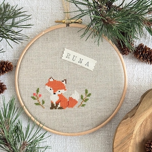 Può includere: Un telaio da ricamo circolare in legno contenente un tessuto beige chiaro. Il tessuto presenta un disegno a punto croce di una piccola volpe arancione e bianca seduta tra due rametti verdi con bacche rosse. Un'etichetta bianca riporta la scritta RUNA.