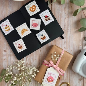 Può includere: Un taccuino nero contenente sette toppe quadrate a punto croce con illustrazioni di cibo e i numeri 60, 10, 2, 25, 5, 3 e 80. Un regalo avvolto in carta marrone presenta un nastro rosa e una toppa a punto croce con il numero 75.