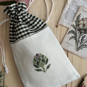 May include: A drawstring pouch made of white fabric with a black and white gingham top section. The front features a small, embroidered green artichoke design. White rope cords are used to close the bag, which is accented by green and pink ribbons.