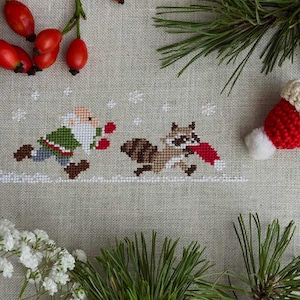 Peut inclure: Une broderie au point de croix sur un tissu en lin beige clair représentant un gnome en manteau vert et un raton laveur courant parmi des flocons de neige brodés en blanc, décoré de cynorhodons rouges et d'aiguilles de pin.