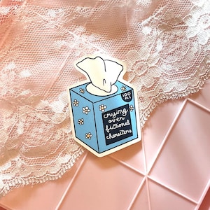 May include: A sticker featuring a blue tissue box with white flowers and the text "crying over fictional characters." The tissue box has a white tissue sticking out. The sticker is on a pink surface with a lace overlay.