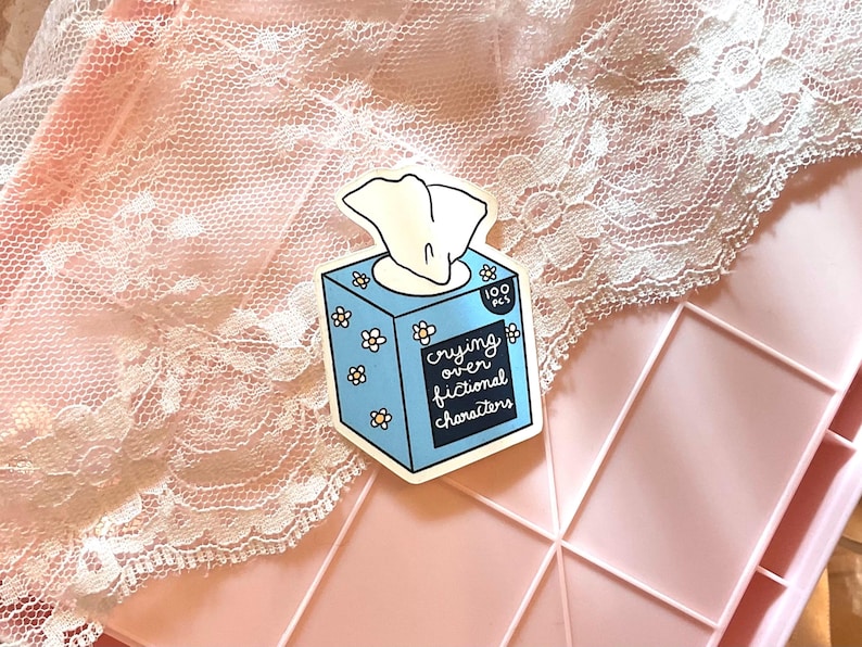 May include: A sticker featuring a blue tissue box with white flowers and the text "crying over fictional characters." The tissue box has a white tissue sticking out. The sticker is on a pink surface with a lace overlay.