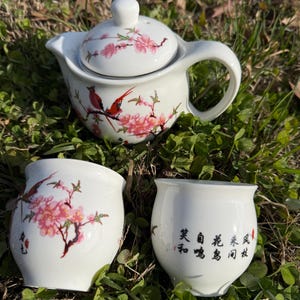 May include: A white ceramic tea set featuring a teapot with a lid and two teacups. The set is decorated with pink cherry blossoms and a red bird. The teacups have Chinese characters. The set is sitting on green grass.