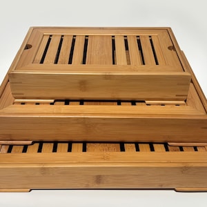 May include: A set of three rectangular bamboo tea trays stacked in descending order of size. Each tray features a light brown natural wood grain, a raised frame, and a slatted surface design with open gaps to allow for drainage during tea service.