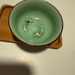 May include: A celadon green ceramic bowl with a dark brown rim, featuring two small koi fish figurines inside. The bowl sits on a small, rectangular bamboo tray. The koi are white, black, and red.