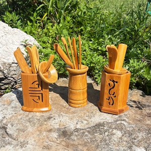 May include: Three bamboo tea sets are displayed outdoors on a rock. Each set includes a container and various tea utensils. The containers have carved designs and hold tools like spoons and tongs. The sets are made of light brown bamboo.