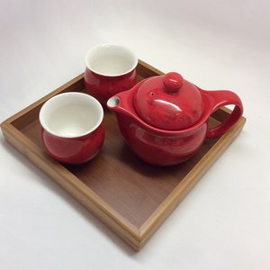May include: A red ceramic teapot and two matching teacups on a wooden tray. The teapot has a lid and a handle. The teacups are small and round.