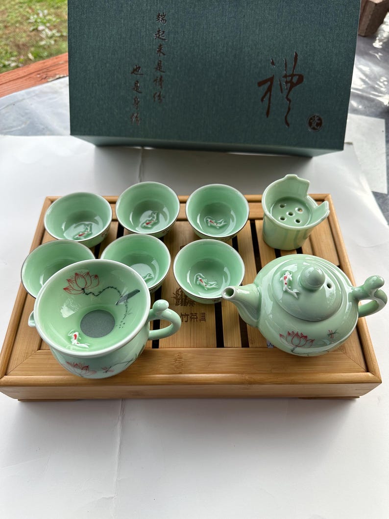 May include: A green tea set with a teapot, cups, and a strainer. The set is decorated with lotus flowers and fish. The set is on a bamboo tray.