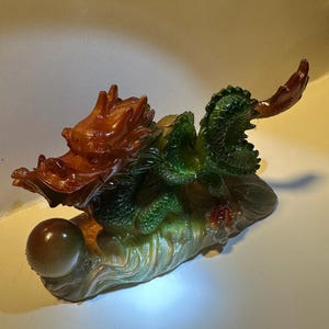 May include: A green and brown dragon figurine holding a pearl. The dragon is perched on a rock with a wave-like design.