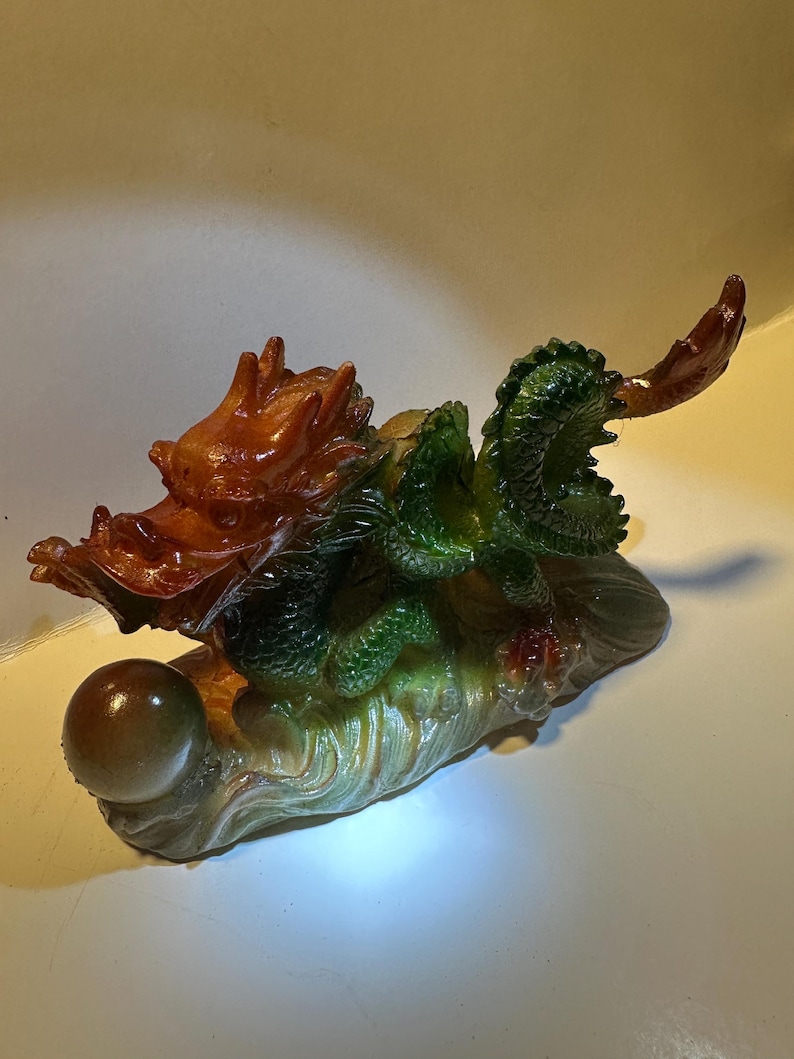 May include: A green and brown dragon figurine holding a pearl. The dragon is perched on a rock with a wave-like design.