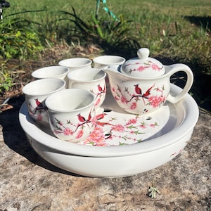 May include: A white ceramic tea set featuring a teapot, six cups, and a serving tray. The set is decorated with pink floral and red bird designs. The tea set is placed on a stone surface.