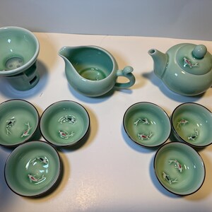 May include: A celadon green tea set featuring a teapot, creamer, strainer, and six small cups. Each cup and the teapot are decorated with small fish. The set is made of ceramic.