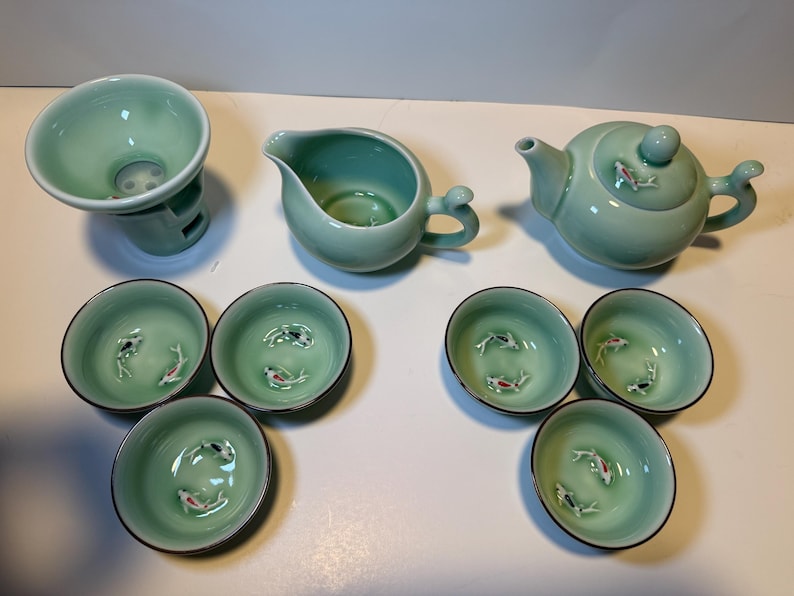 May include: A celadon green tea set featuring a teapot, creamer, strainer, and six small cups. Each cup and the teapot are decorated with small fish. The set is made of ceramic.